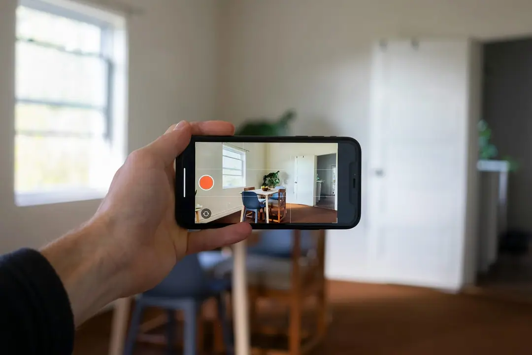 Ranka telefonu fotografuoja šviesų kambarį su stalu ir kėdėmis